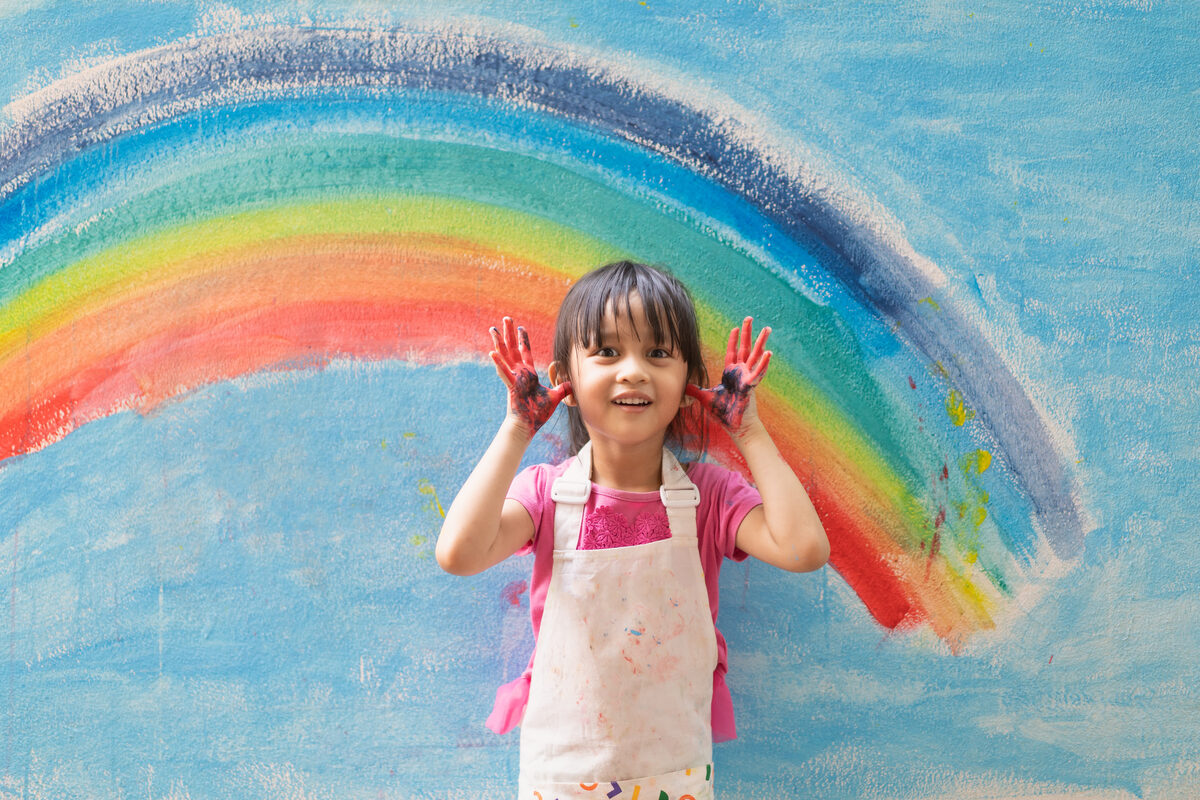 A child smiling proudly in front of a rainbow she painted — a moment shared by a nanny